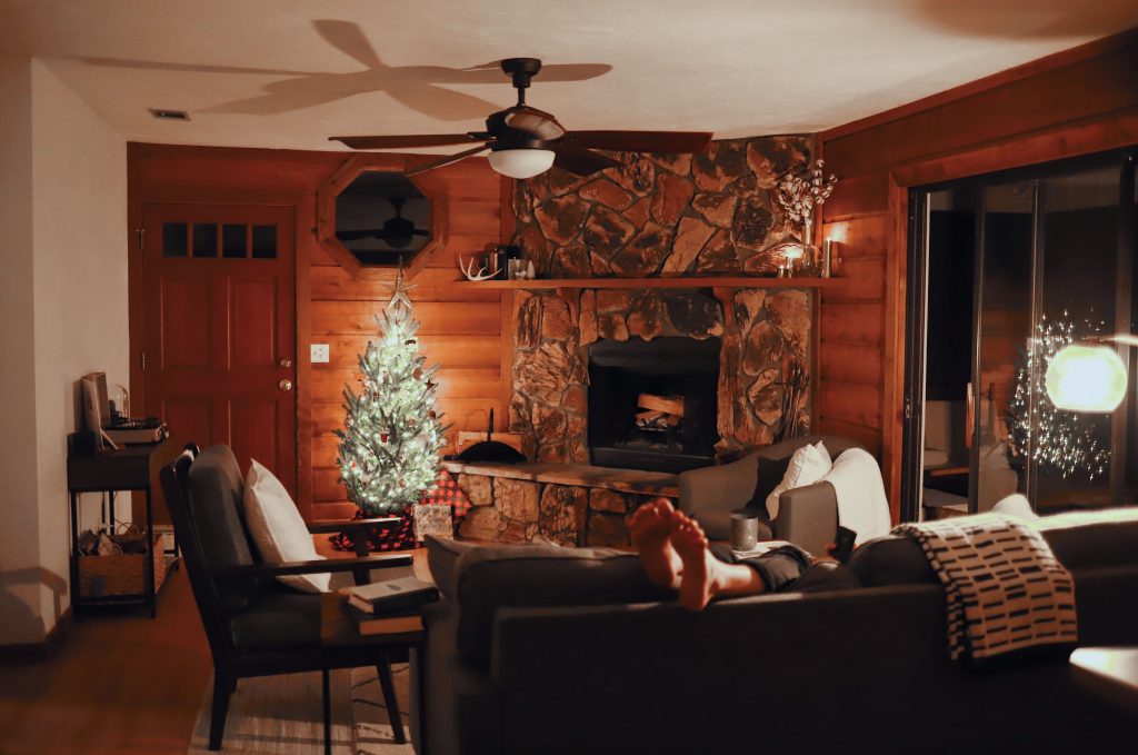 A cozy Christmas at home featuring the feet of a man draped over his couch with comfy throw blankets and a lit up Christmas tree by a fireplace in a wood paneled room.