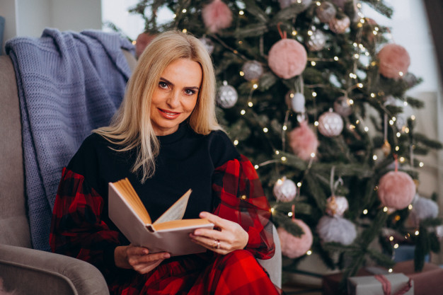 A woman sits neatly groomed and dressed up for Christmas in a comfy chair holding a book with a Christmas tree behind her. 