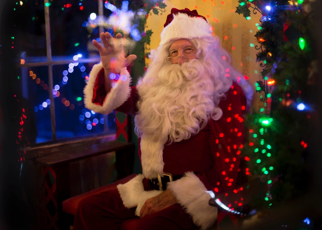 Santa Clause sits in front of a Christmas Tree waving.