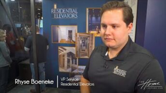 Residential Elevators create Accessible Home, elevator showroom with dark blue walls, photos of construction, man in black polo and short dark hair. Text: Rhyse Boeneke, VP Service, Residential Elevators