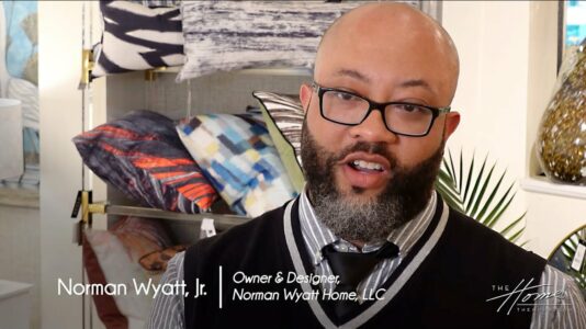 Norman Wyatt Home Lifestyle Collection with StyleCraft showroom, black man with dark beard and black-rimmed glasses in black and white vest, colorful throw pillows in background. Text: Norman Wyatt, Jr., Owner & Designer, Norman Wyatt Home, LLC
