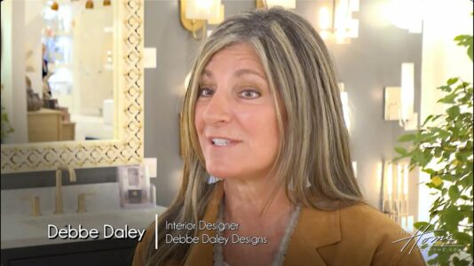 Debbe Daley with long blonde hair and leather blazer and white dress in interior design High Point Market Currey & Company lighting showroom