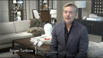 Roger Turnbow, president of A.R.T. Home Furniture sits in gray wool suit with gray hair and beard, in a High Point Market furniture showroom of white couch and dark furniture behind him