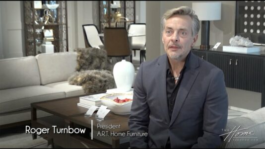 Roger Turnbow, president of A.R.T. Home Furniture sits in gray wool suit with gray hair and beard, in a High Point Market furniture showroom of white couch and dark furniture behind him