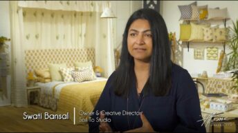 Indian woman with long black hair and black shirt, Swati Bansal, owner & creative director of New York based Soil To Studio textile design studio in High Point Market showroom with colorful bedding, pillows and fabrics behind her