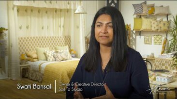 Indian woman with long black hair and black shirt, Swati Bansal, owner & creative director of New York based Soil To Studio textile design studio in High Point Market showroom with colorful bedding, pillows and fabrics behind her