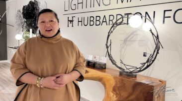 Designer Jeanne Chung in tan dress stands next to a modern wrought iron lamp and wooden table in Hubbardton Forge showroom