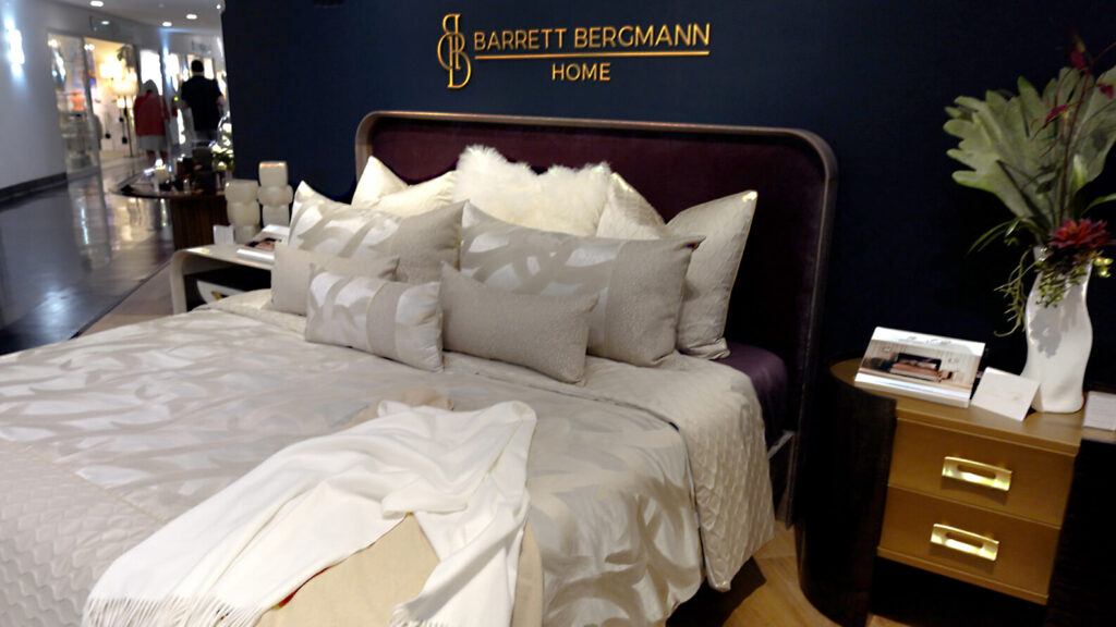 Image: dark Barrett Bergmann Home showroom with taupe bedding and pillows and white furry pillow, nightstand with floral arrangement