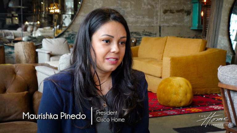 Image: Latino woman with navy jacket in furniture showcase room in front of a curry brown sofa, red rug, ottoman, and brown leather chair