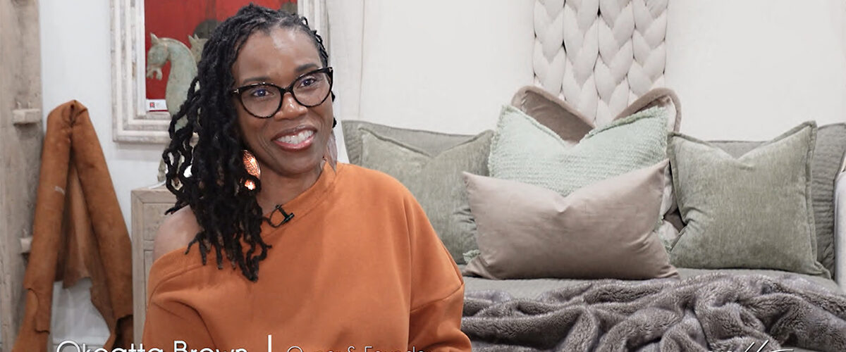 Image of smiling black woman with black rim glasses and braided hair wearing orange sweater on foot of Victoria Lane Collection gray bed with white braided headboard