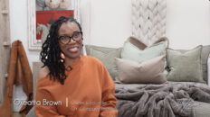 Image of smiling black woman with black rim glasses and braided hair wearing orange sweater on foot of Victoria Lane Collection gray bed with white braided headboard