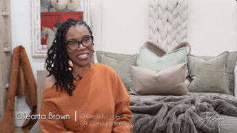 Image of smiling black woman with black rim glasses and braided hair wearing orange sweater on foot of Victoria Lane Collection gray bed with white braided headboard