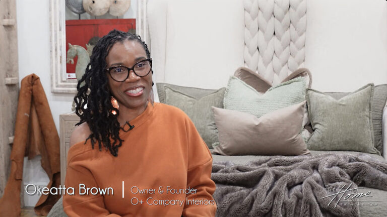 Image of smiling black woman with black rim glasses and braided hair wearing orange sweater on foot of Victoria Lane Collection gray bed with white braided headboard