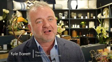 Kyle Barrett of Barrett Bergmann Home wearing gray jacket in home furnishings and decor showroom with Ikebana silk floral arrangements and pillows in background