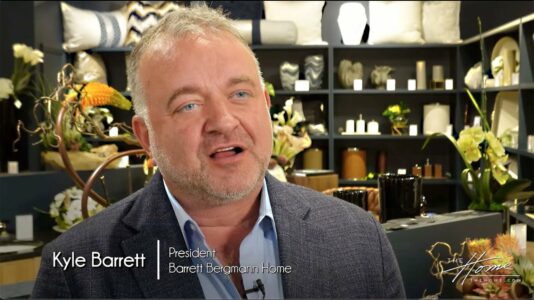Kyle Barrett of Barrett Bergmann Home wearing gray jacket in home furnishings and decor showroom with Ikebana silk floral arrangements and pillows in background