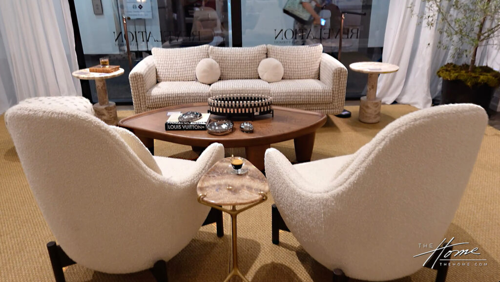 Image: furniture showroom with off-white checkered couch w/ 2 round upholstered offwhite pillows, organic shaped wood coffee table, 2 textured off-white upholstered chairs with accent marble side table, jute rug