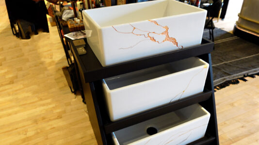 Image: 3 white Thompson Traders Kintsugi fireclay apron sinks with cracks filled with copper, gold and silver on a black rack in showroom