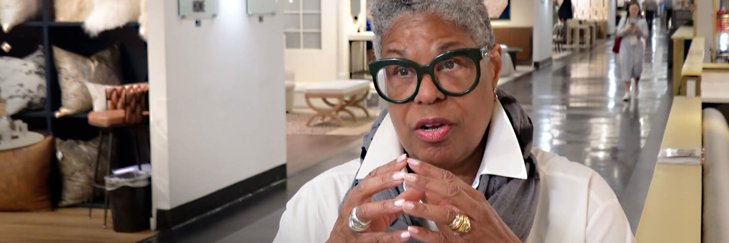 Image: black woman with short gray hair and black rimmed glasses in white shirt in High Point Market showroom aisle
