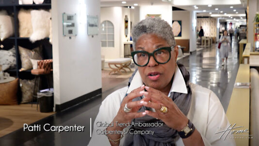Image: black woman with short gray hair and black rimmed glasses in white shirt in High Point Market showroom aisle