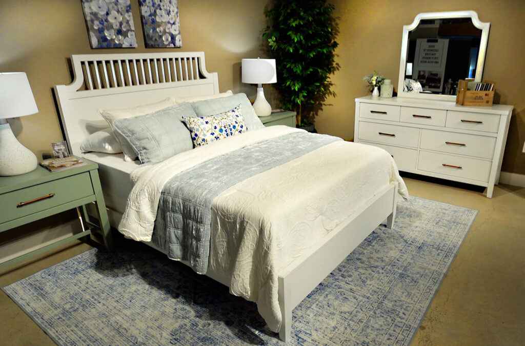 Erin & Ben Napier designed white wood bed headboard with cutout and slat design, white and blue bedding on blue rug, white dresser and mirror in furniture showroom