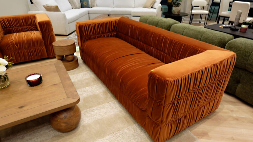 Jessica Simpson Home Collection featured Burnt orange/rust colored velvet sofa with pleats in furniture showroom by wood coffeetable with large sphere legs and green velvet sofa