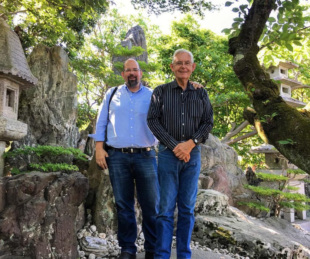 man with beard and blue shirt standing next to older man with white hair and striped shirt in beautiful bonzai garden