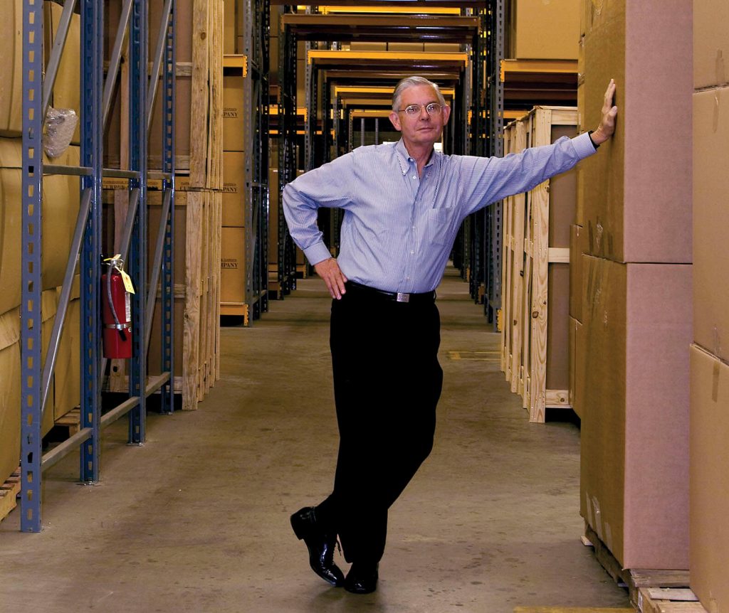 Robert Currey of Currey & Company in button down blue shirt and black dress slacks leans on boxes in warehouse